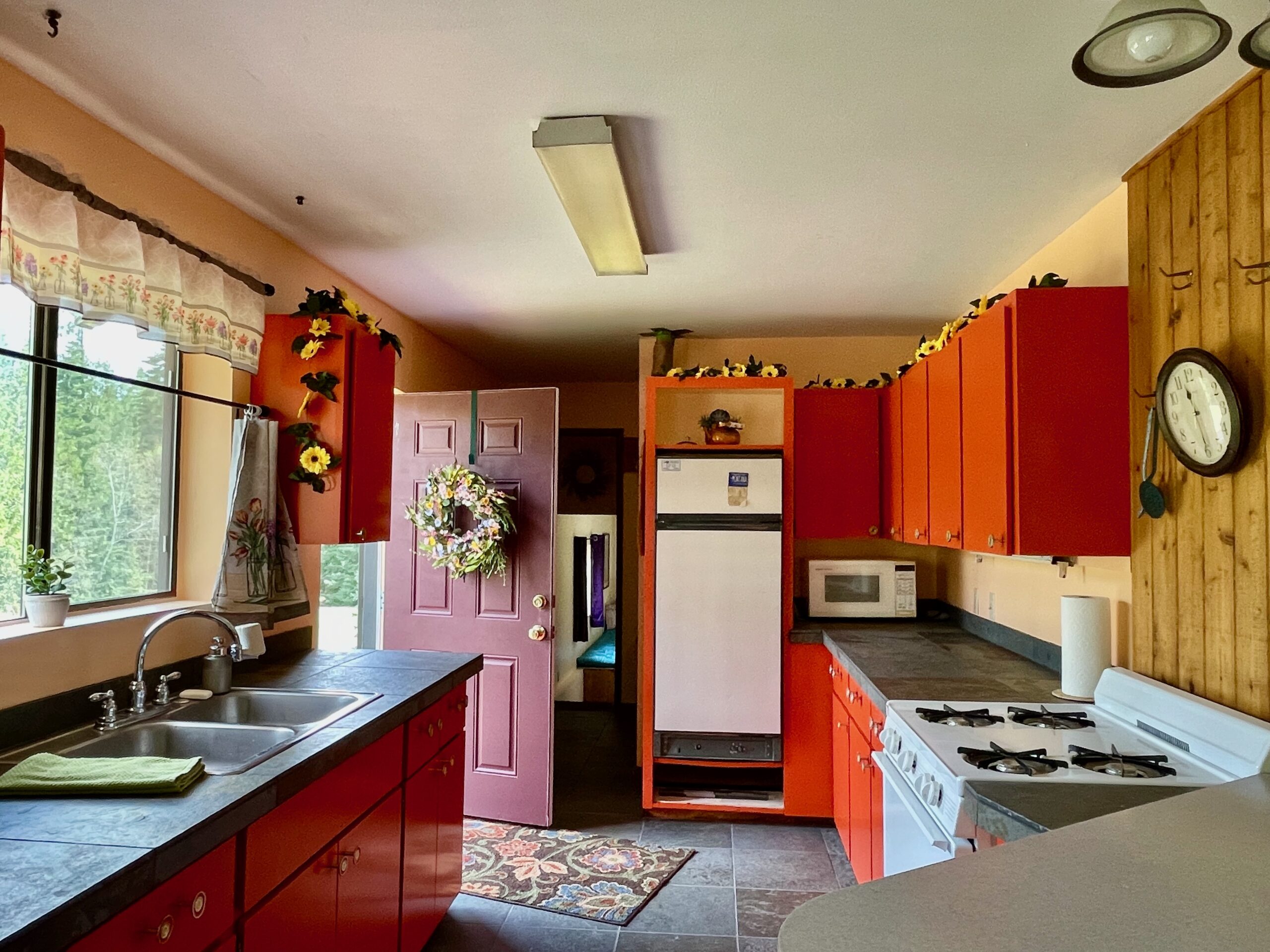 Open front door leading into a peach colored kitchen with warm fiesty orange cabinets and tile countertops