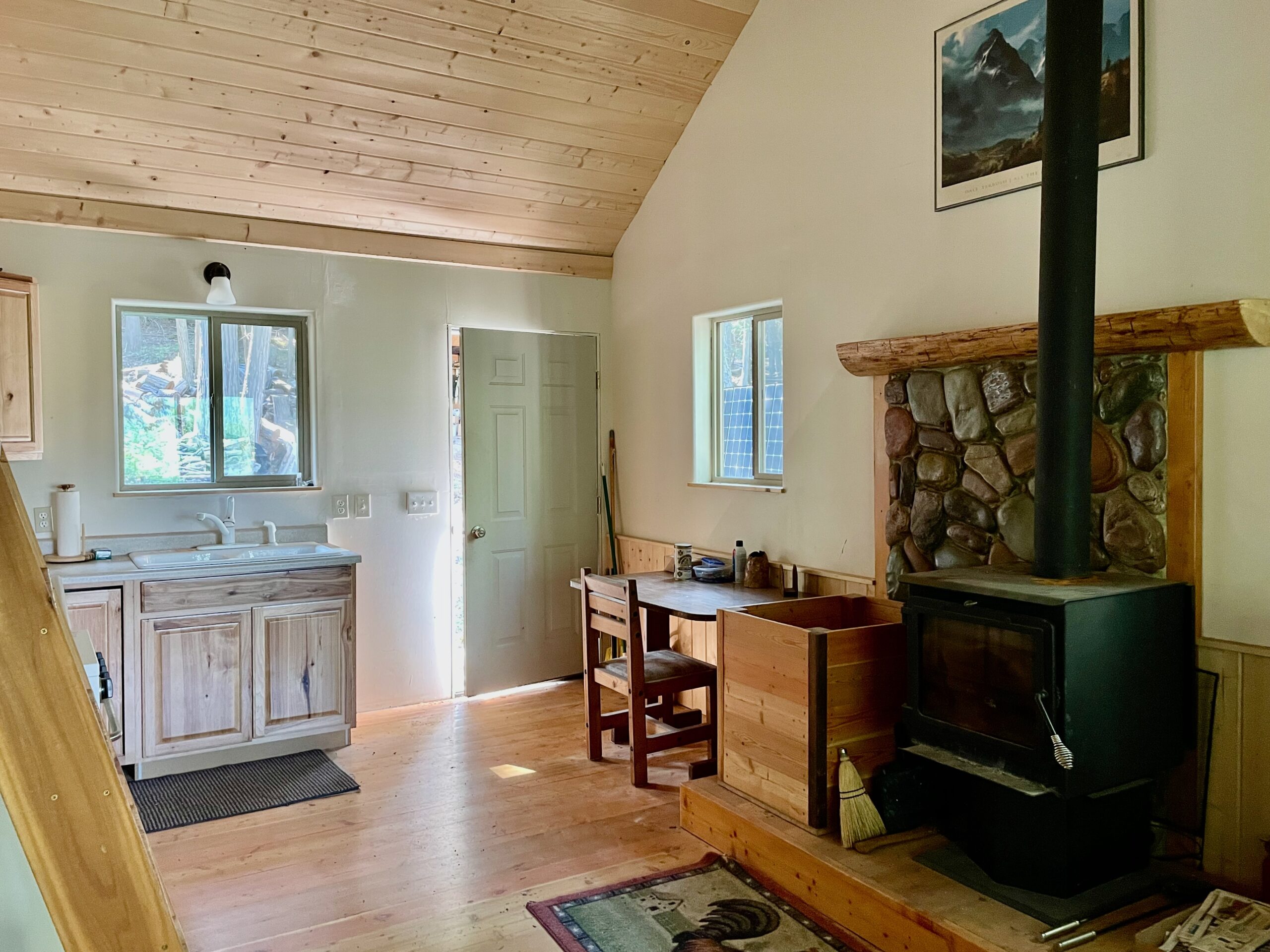 Looking toward the front door, the wood stove with river rock wall behind it is on the right the kitchen sink with a window above it are to the left of the front door.