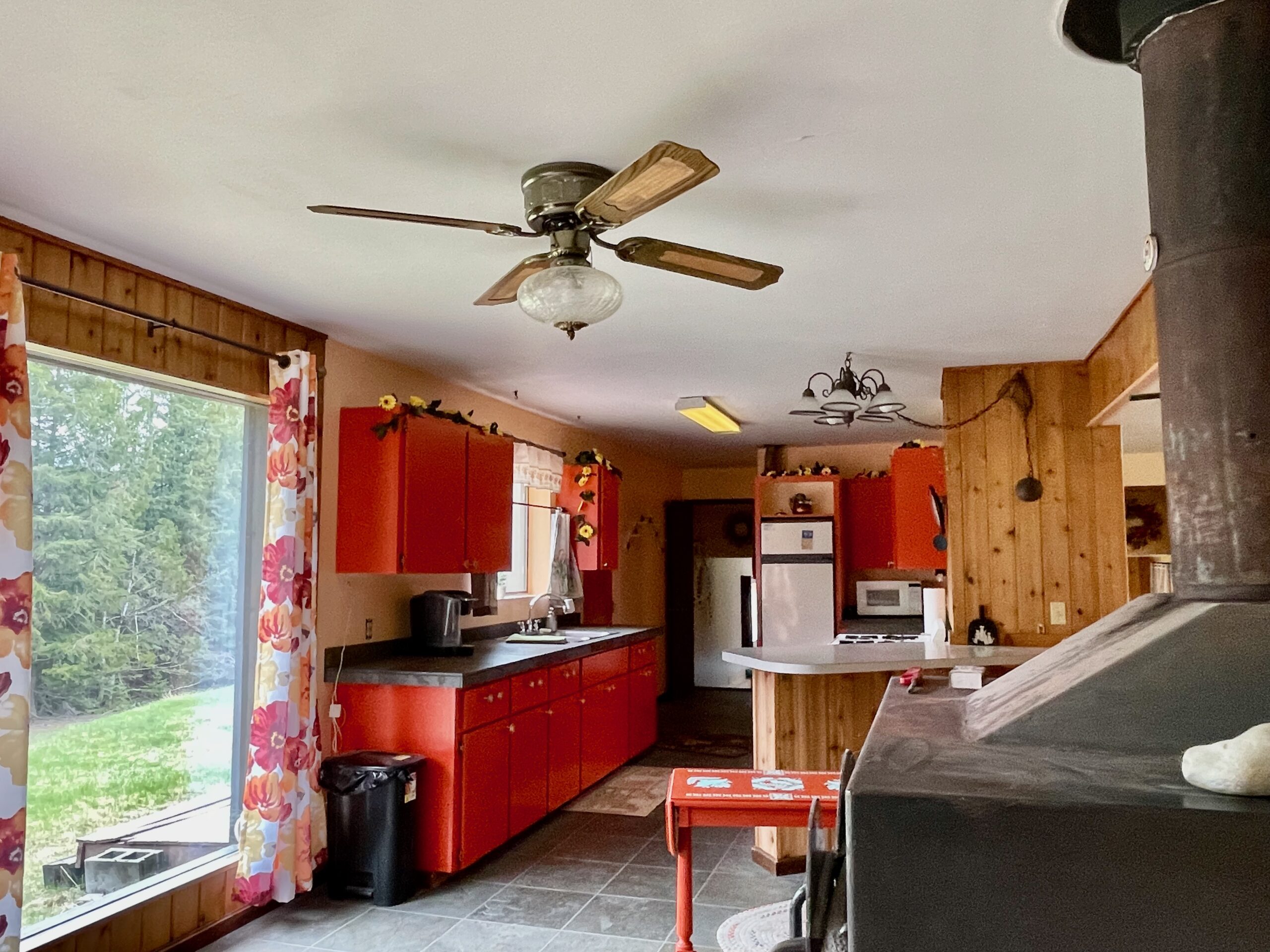 Another view of the large woodstove on the right and a peek out the front window at the lawn and pine trees. The kitchen is in the background. 