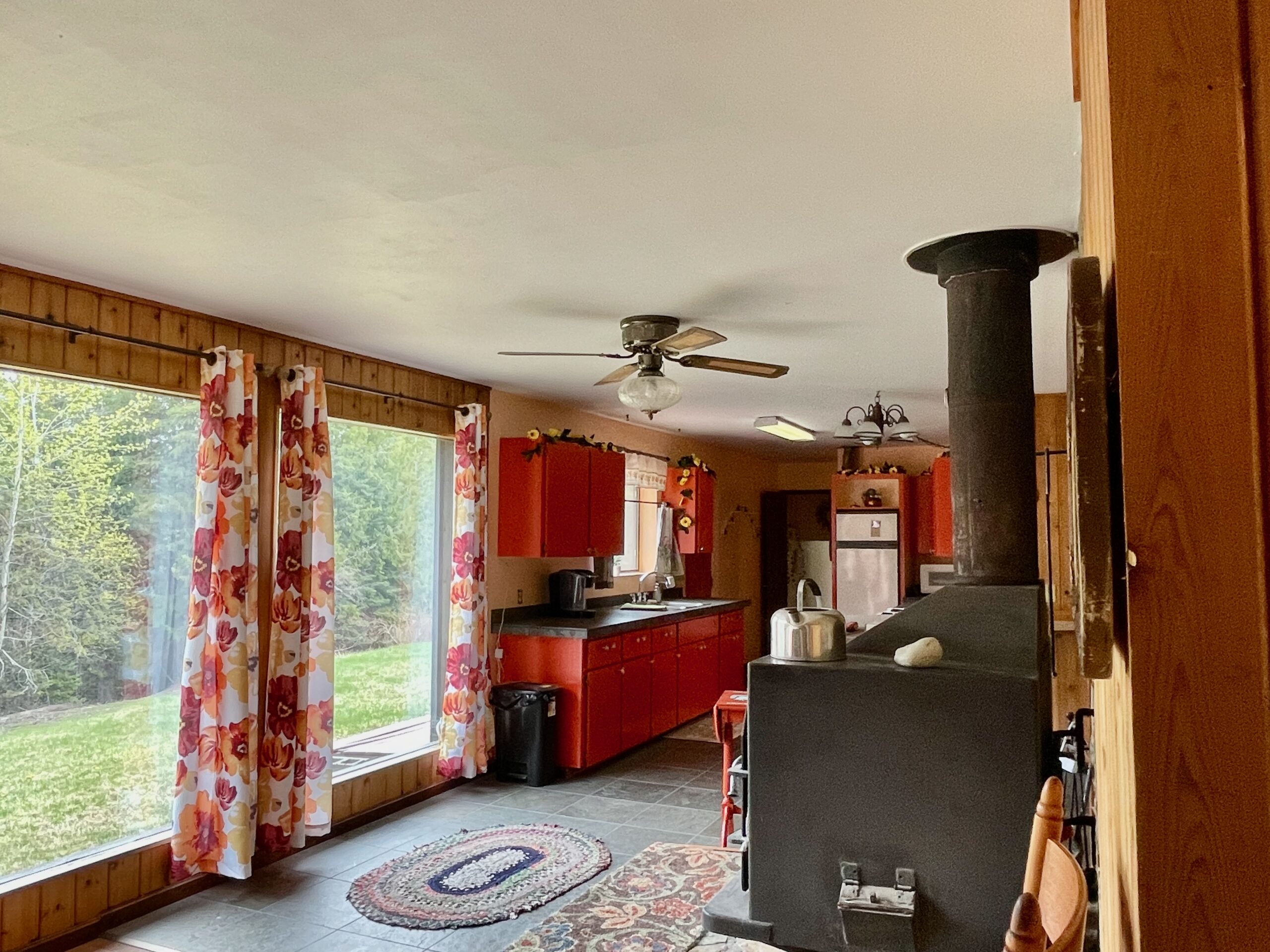 Coming back out of the living room there are the large front windows on the left and the large wood stove on the right and the kitchen in the background.
