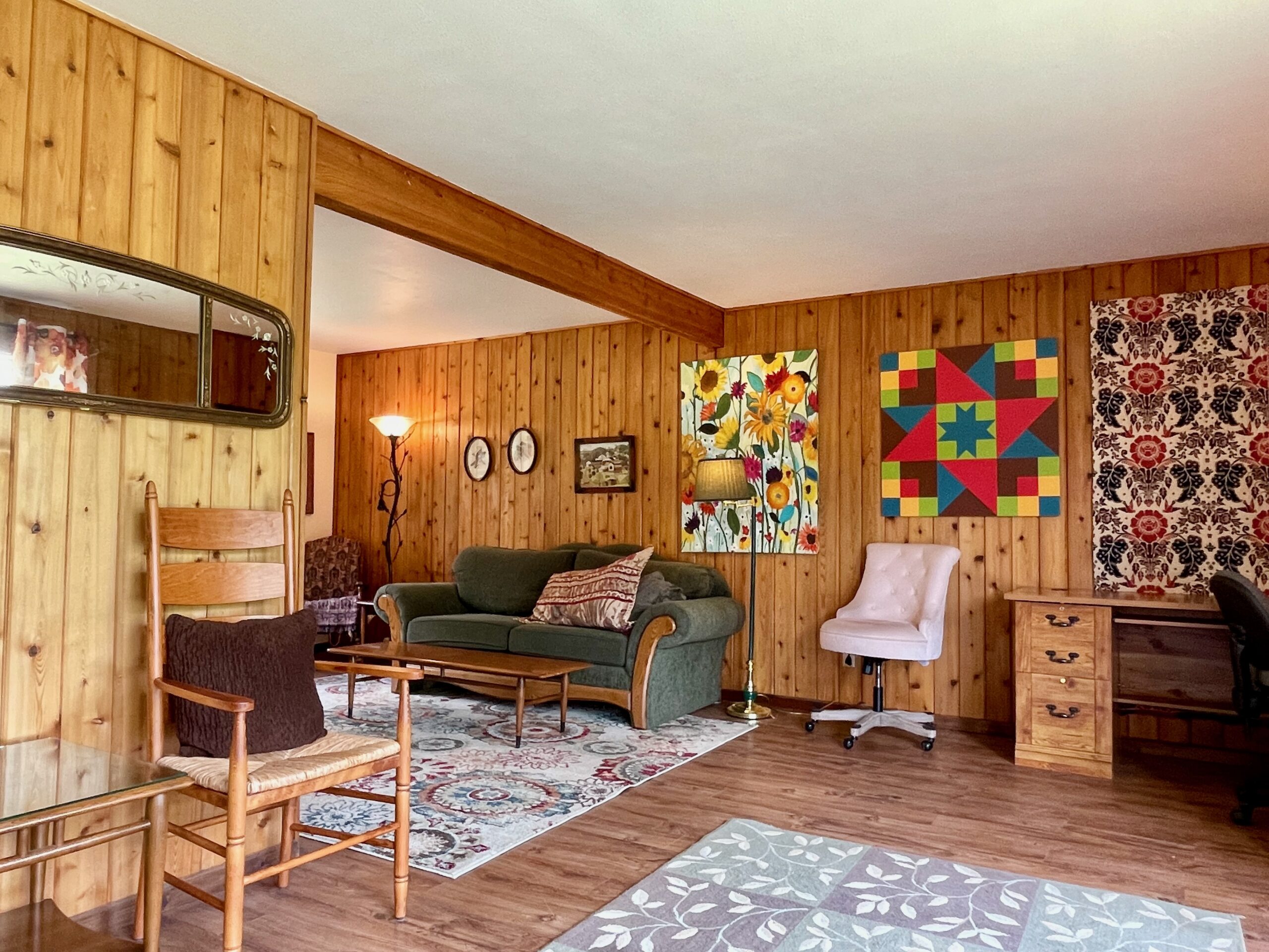 Wood paneling in living room with a corner desk and various types of artwork on walls.